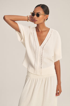 A woman wearing sunglasses and gold hoop earrings pairs the Velvet by Graham & Spencer VERNA TOP—a white cotton gauze top with lace cutout details and a deep V-neck—with a matching white skirt.