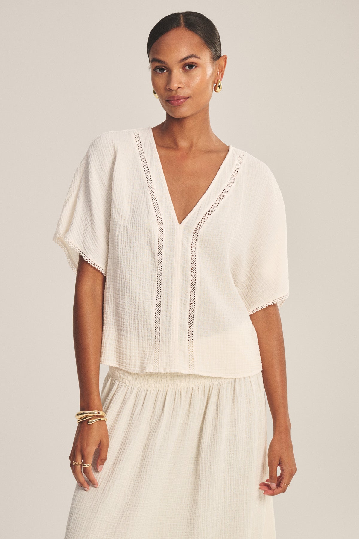A woman wears the Velvet by Graham & Spencer VERNA TOP, a white, short-sleeved cotton gauze blouse with subtle lace trim, paired with a matching skirt and gold earrings and bracelets.