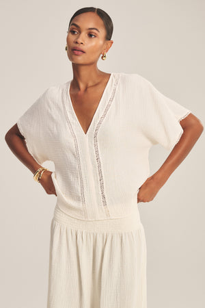 A woman wears the VERNA TOP by Velvet by Graham & Spencer, a loose-fitting, short-sleeve cotton gauze top with lace cutout details, paired with a matching white skirt and standing with hands on hips against a neutral background.