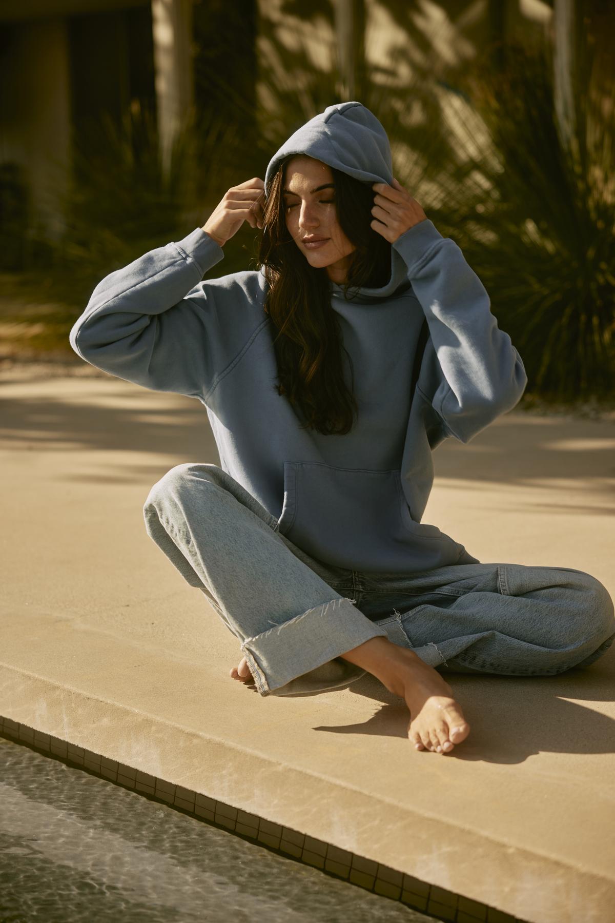   A person in a VERONICA HOODIE by Velvet by Graham & Spencer, paired with jeans, sits cross-legged outdoors by a pool, adjusting the hood while enjoying the sunlight. 