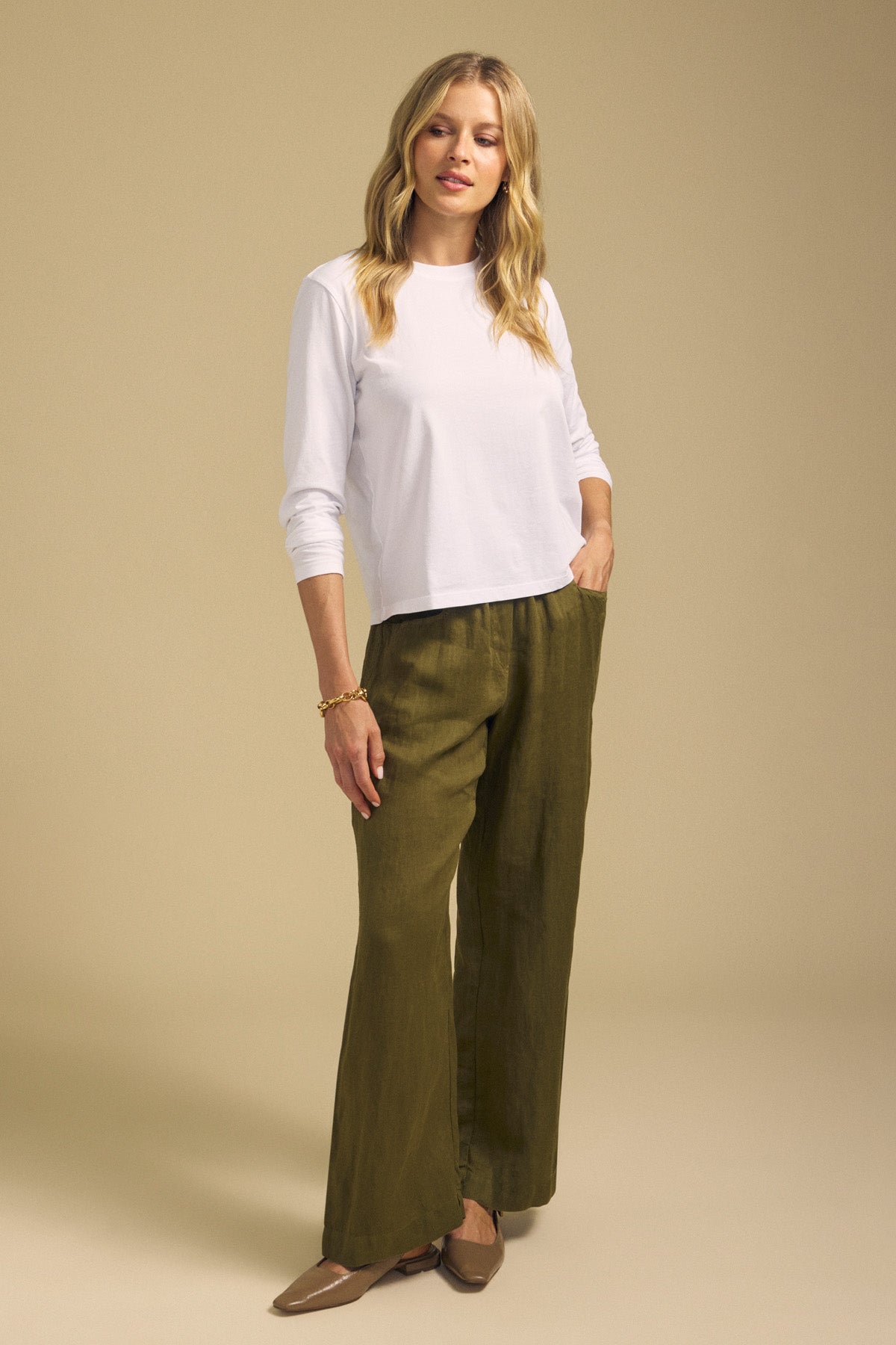 A person stands facing forward against a plain beige background, wearing a long-sleeve white shirt and CLAIRO LINEN PANT by Velvet by Graham & Spencer—an olive green, lightweight linen pant featuring a pull-on elastic waist and wide-leg silhouette—paired with brown flats and a bracelet.