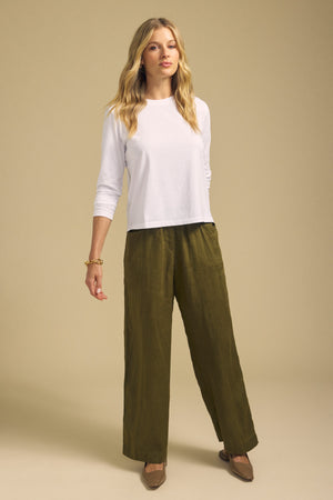 A person stands against a beige background wearing the VICKY TEE by Velvet by Graham & Spencer, a white long sleeve cotton jersey top, paired with olive green wide-leg cropped pants, brown flats, and a bracelet.