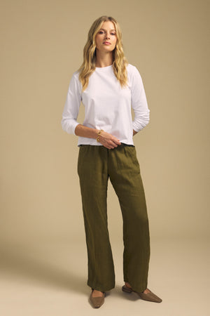 A woman stands against a beige background wearing a white long-sleeve shirt, brown flats, a bracelet, and the CLAIRO LINEN PANT from Velvet by Graham & Spencer—an olive green pull-on style featuring an elastic waist and wide-leg silhouette.