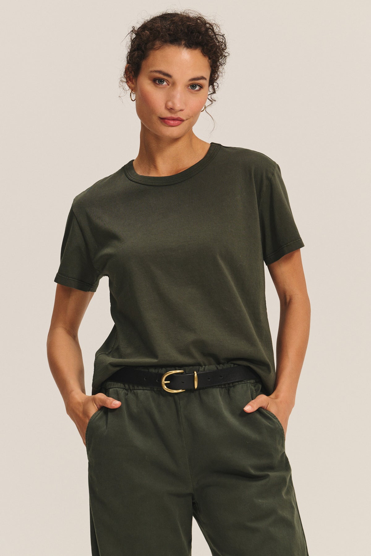   A woman stands with hands in pockets against a neutral background, wearing the LEIGH TEE by Velvet by Graham & Spencer—a relaxed fit, olive green cotton jersey t-shirt—tucked into matching pants with a black belt. 