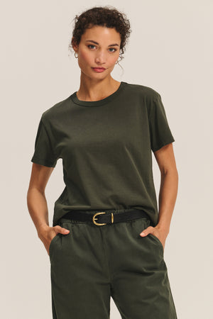 A woman stands with hands in pockets against a neutral background, wearing the LEIGH TEE by Velvet by Graham & Spencer—a relaxed fit, olive green cotton jersey t-shirt—tucked into matching pants with a black belt.