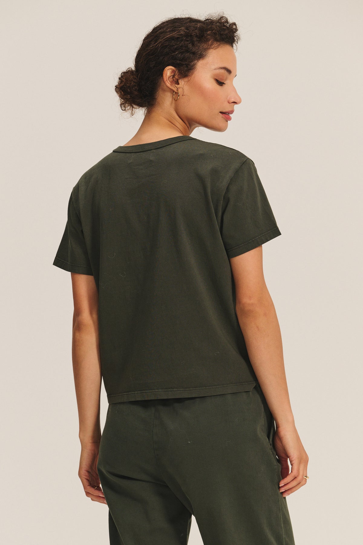 A person stands with their back to the camera, wearing the LEIGH TEE by Velvet by Graham & Spencer in dark green cotton jersey, paired with matching pants, hair tied back, against a plain light background.-40163404972225
