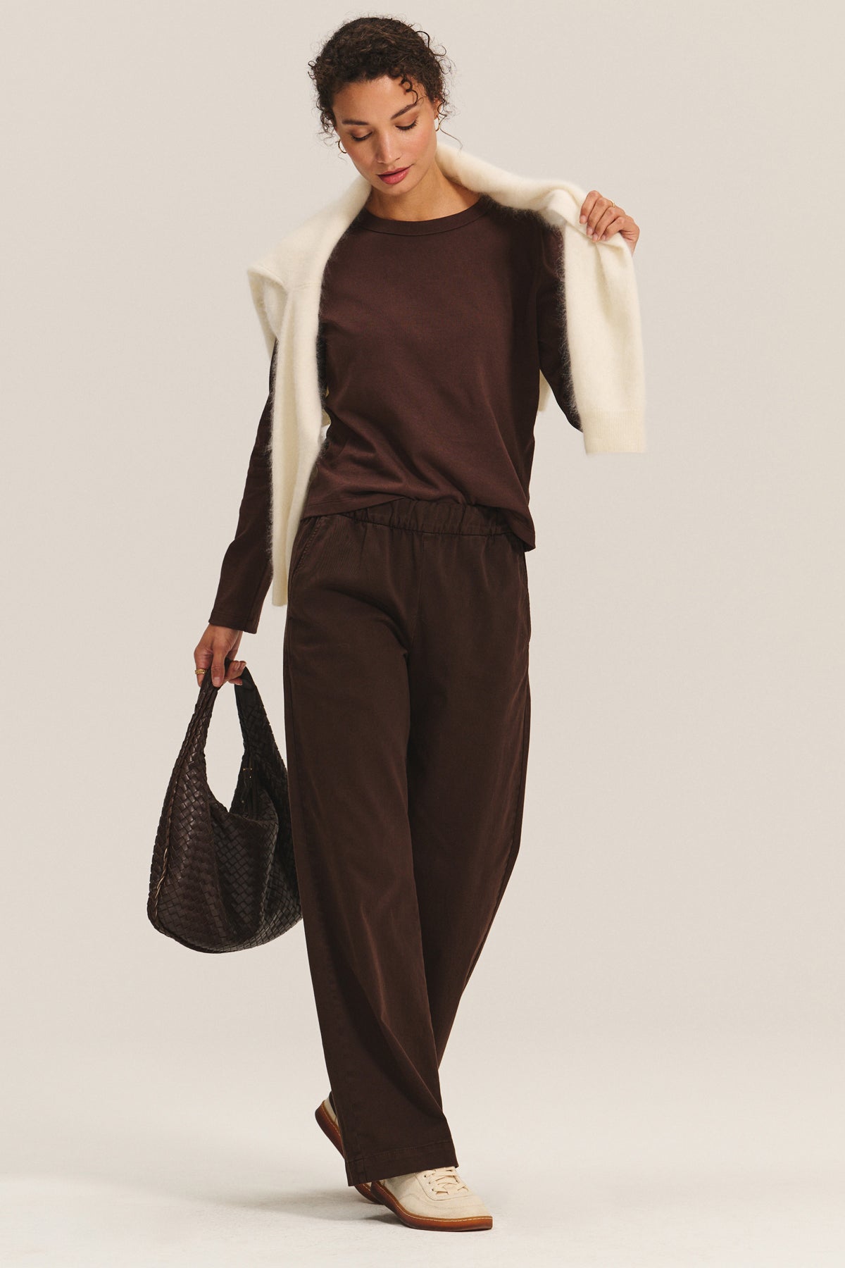   A person wears the SUSANNA TEE by Velvet by Graham & Spencer—vintage-inspired brown long-sleeve top—with matching brown pants, a cream sweater over the shoulders, cream sneakers, and a textured brown bag. 