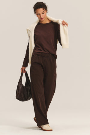 A person wears the SUSANNA TEE by Velvet by Graham & Spencer—vintage-inspired brown long-sleeve top—with matching brown pants, a cream sweater over the shoulders, cream sneakers, and a textured brown bag.