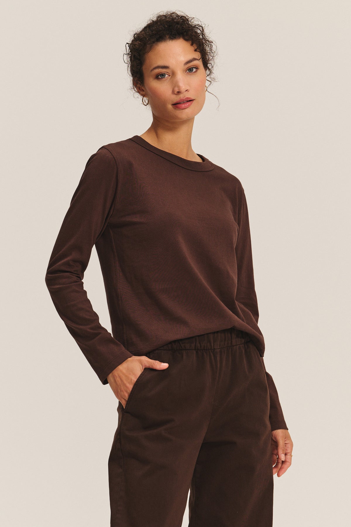 A person models the SUSANNA TEE by Velvet by Graham & Spencer— a vintage-inspired, long-sleeve brown cotton jersey shirt—paired with brown pants, standing with one hand in their pocket against a neutral background.-40182501867713