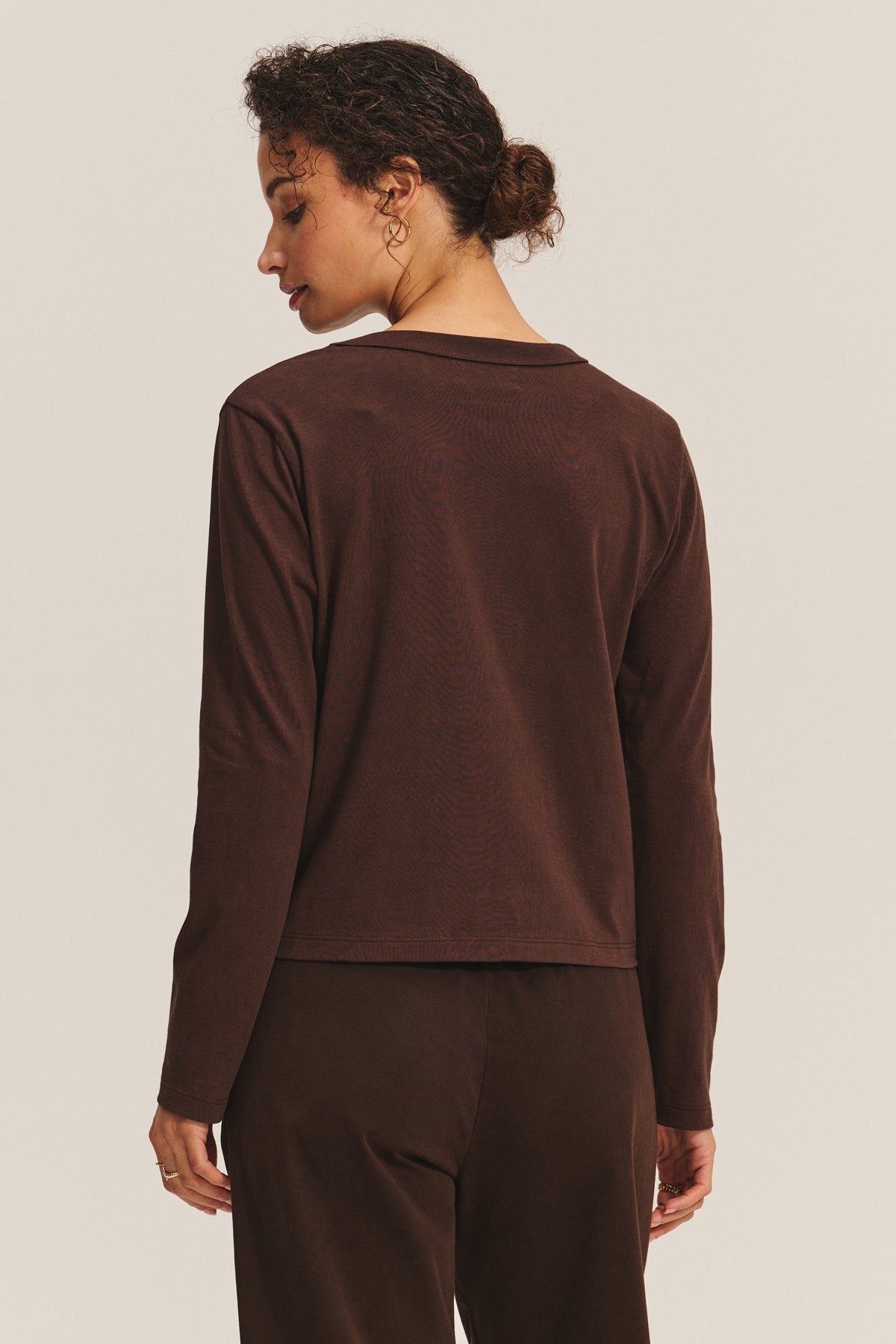   A person with curly hair in a low bun faces away from the camera, wearing the SUSANNA TEE by Velvet by Graham & Spencer—a long-sleeve, vintage-inspired dark brown cotton jersey top—paired with matching pants against a plain background. 