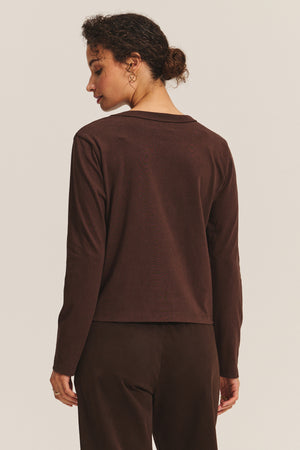 A person with curly hair in a low bun faces away from the camera, wearing the SUSANNA TEE by Velvet by Graham & Spencer—a long-sleeve, vintage-inspired dark brown cotton jersey top—paired with matching pants against a plain background.