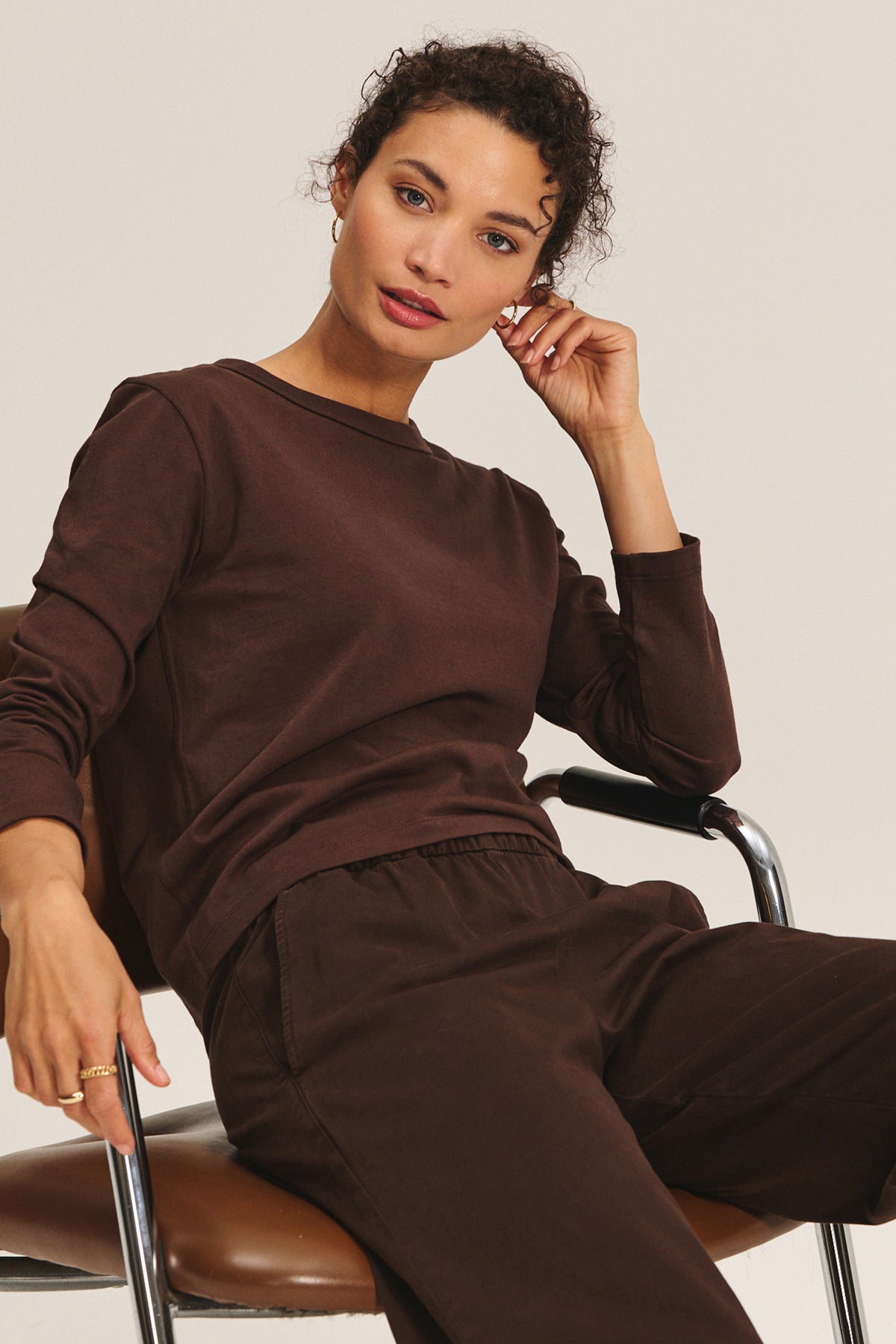   A person with curly hair sits on a chair, wearing the SUSANNA TEE by Velvet by Graham & Spencer in brown and matching pants, resting their left elbow on the chair arm while looking toward the camera. 