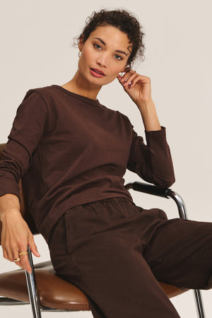 A person with curly hair sits on a chair, wearing the SUSANNA TEE by Velvet by Graham & Spencer in brown and matching pants, resting their left elbow on the chair arm while looking toward the camera.