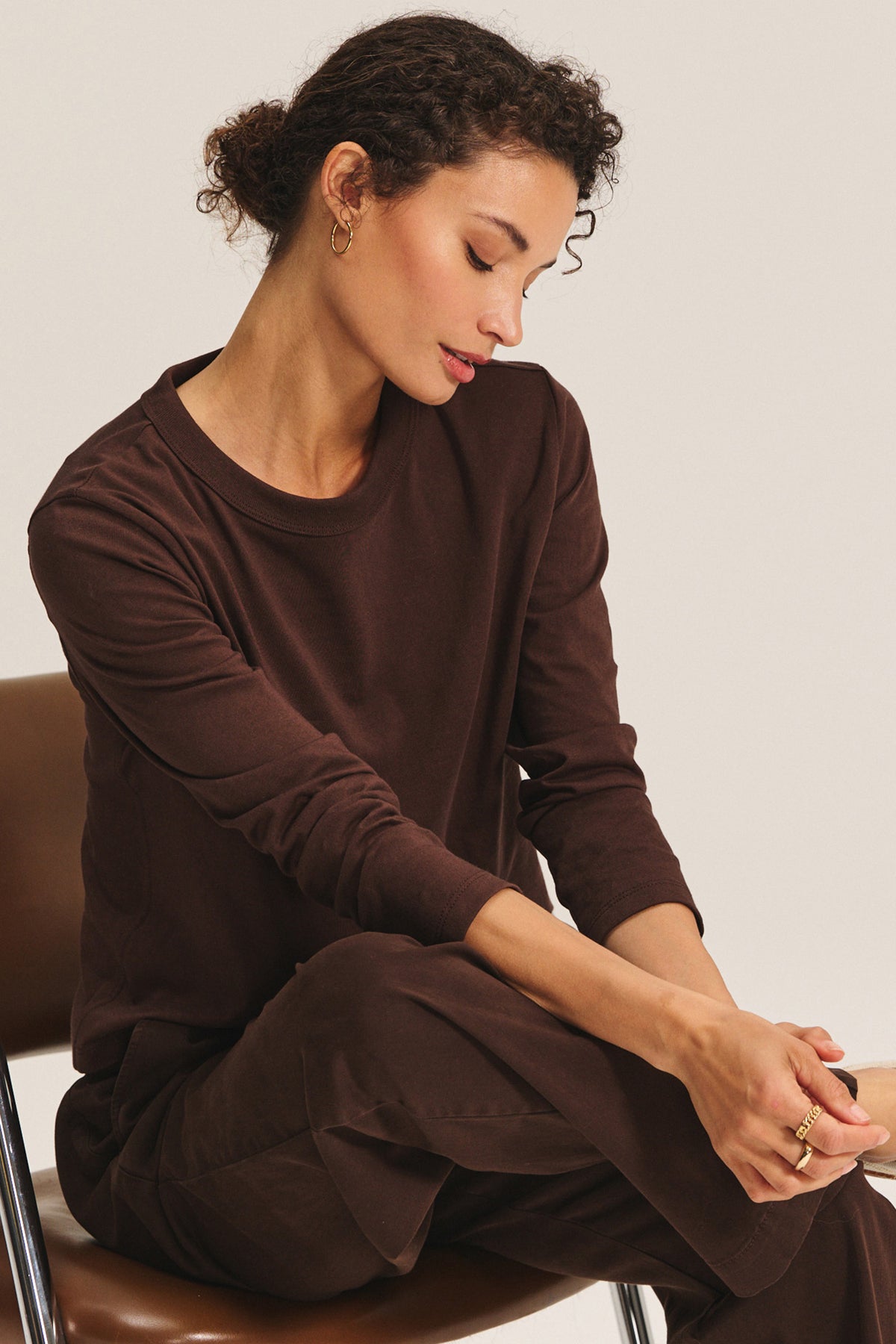   A woman with curly hair sits on a chair, looking down with her hands clasped, wearing the Velvet by Graham & Spencer SUSANNA TEE and brown pants. 