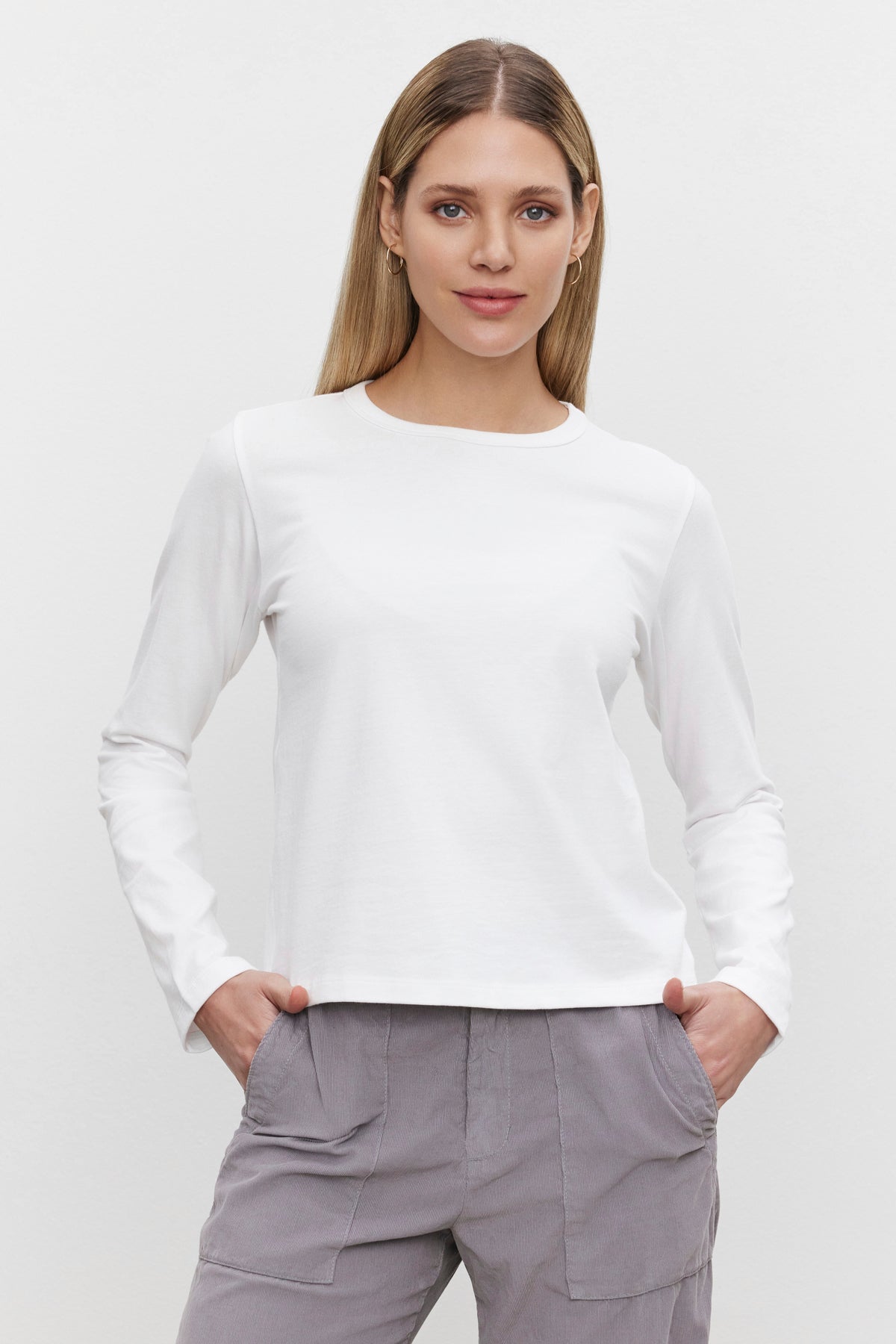   A woman, dressed in the SUSANNA TEE from Velvet by Graham & Spencer—a white long-sleeve shirt with a classic crew neckline—and gray pants, stands against a plain white background with her hands in her pockets. 