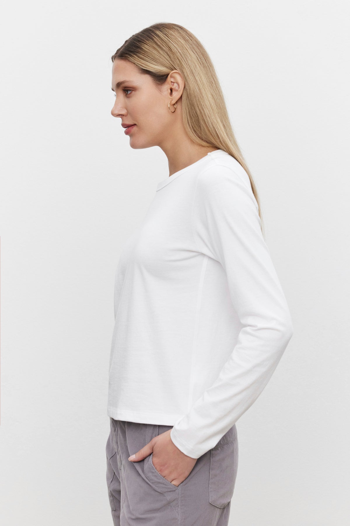   A person with long blonde hair is shown in profile, wearing a vintage-inspired look featuring the SUSANNA TEE by Velvet by Graham & Spencer—a white long-sleeve shirt with a classic crew neckline—and light gray pants, all set against a plain, white background. 