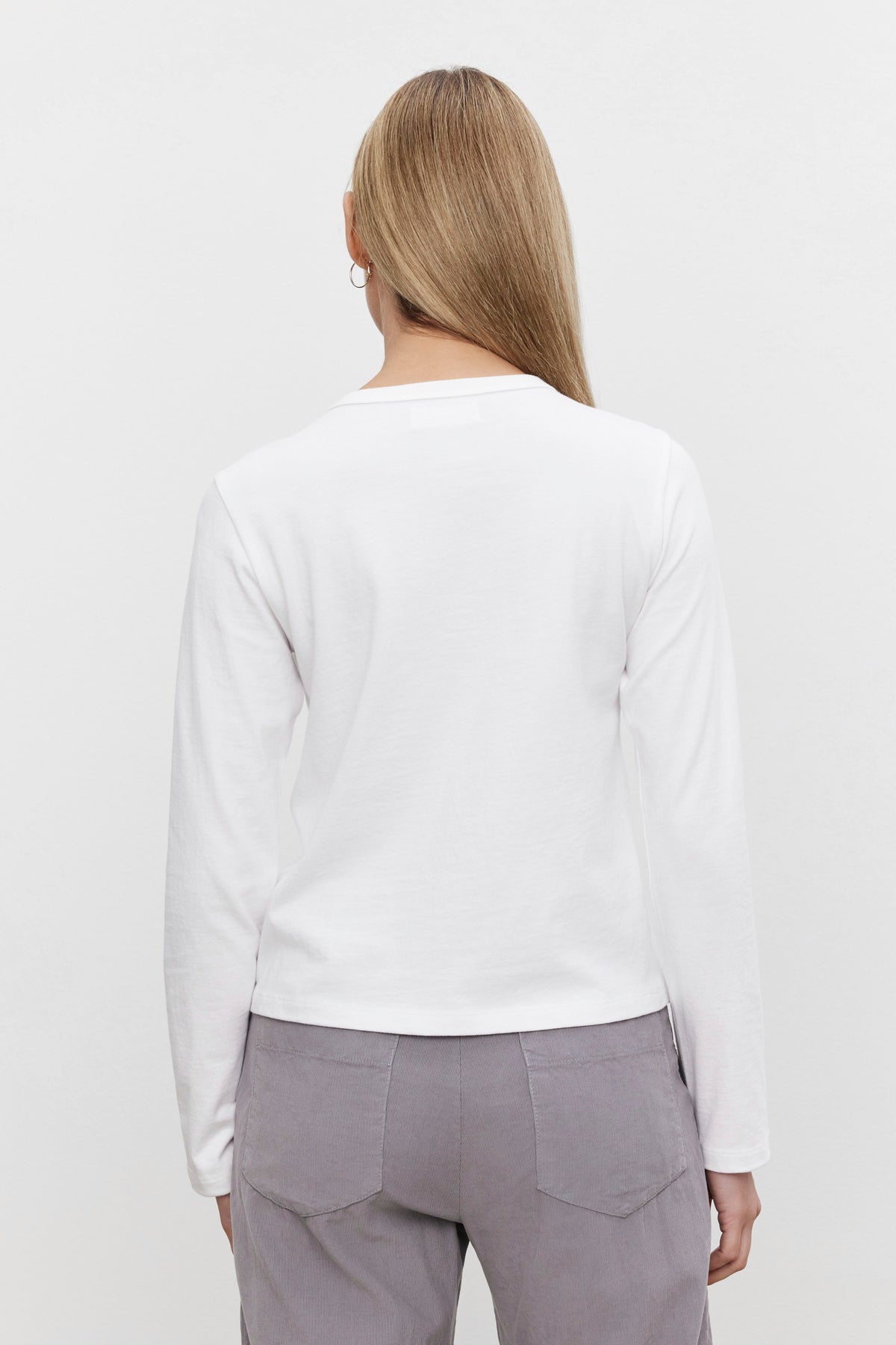   A person with long hair is shown from the back wearing a white, long-sleeved SUSANNA TEE by Velvet by Graham & Spencer featuring a classic crew neckline, paired with gray pants, creating a vintage-inspired look against a plain background. 