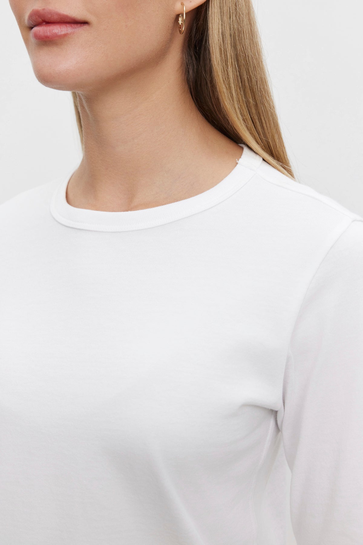   A person wearing the SUSANNA TEE, a vintage-inspired white long-sleeved shirt from Velvet by Graham & Spencer, with a classic crew neckline and a gold hoop earring. The image captures the person's neck and partial face. 