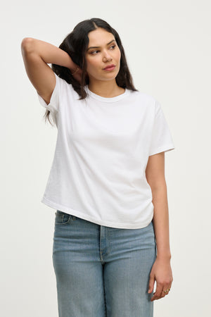 A woman with long dark hair wears the relaxed fit LEIGH TEE by Velvet by Graham & Spencer and blue jeans, standing with one hand behind her head and looking to the side against a plain background.