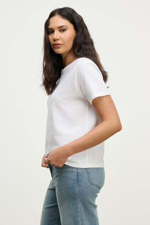 A woman with long dark hair wears the LEIGH TEE by Velvet by Graham & Spencer, a relaxed fit white cotton jersey t-shirt, paired with light blue jeans against a plain light background.