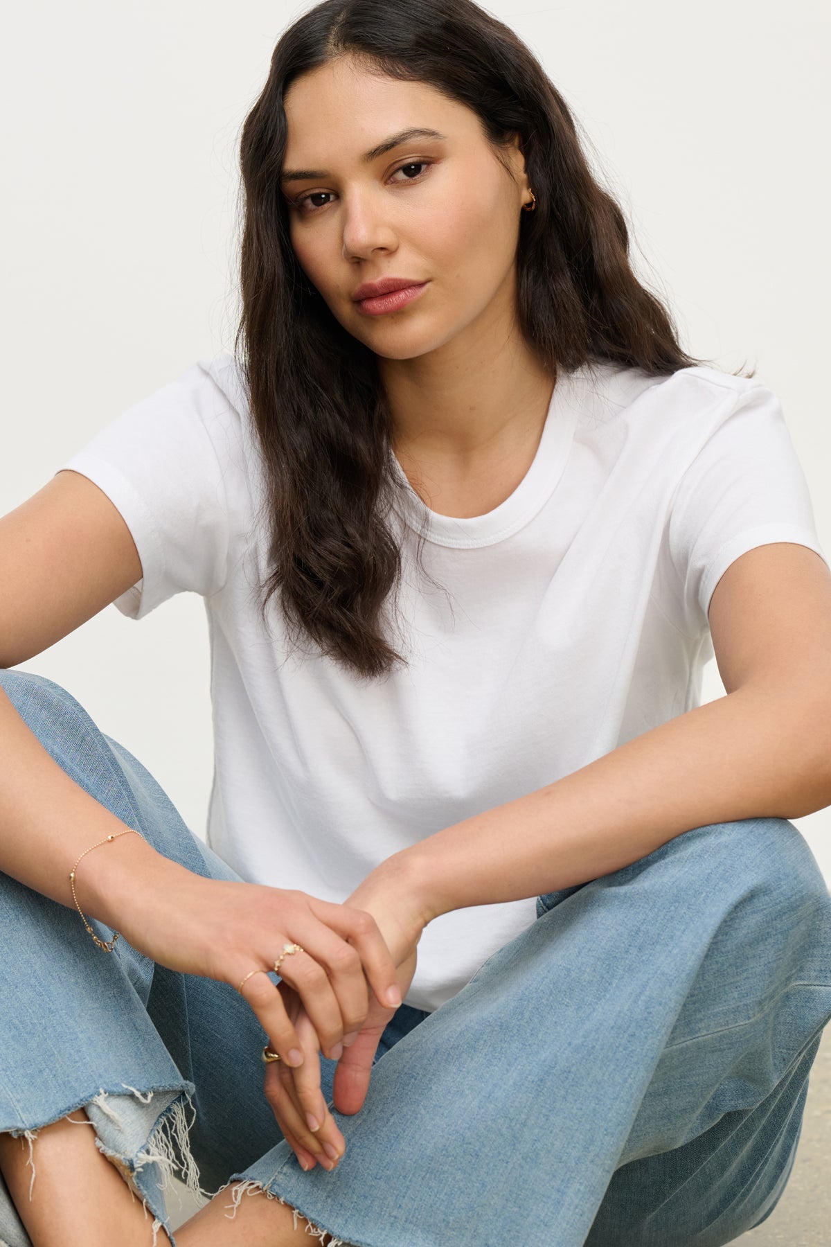 A woman with long dark hair sits cross-legged against a plain background, wearing the LEIGH TEE by Velvet by Graham & Spencer and light blue jeans.-39742241505473