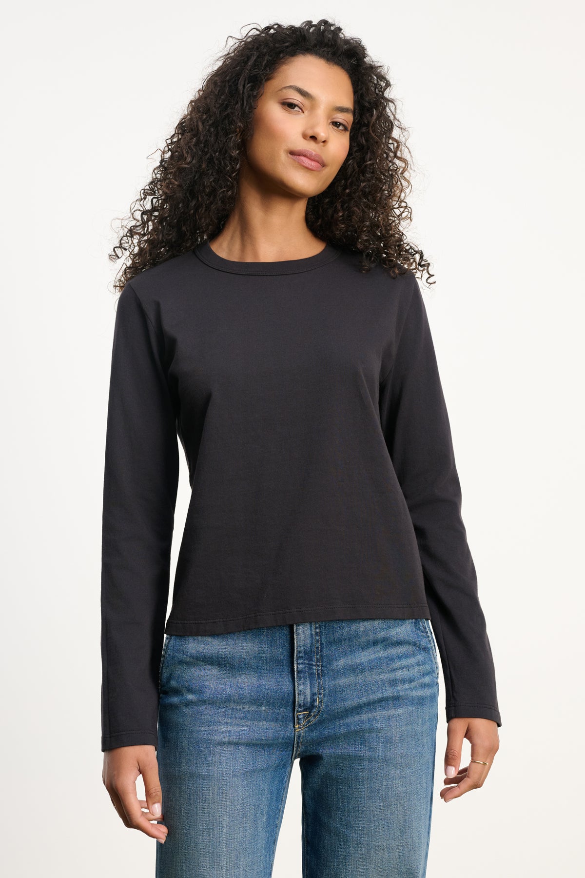 A woman with curly hair wears the SUSANNA TEE by Velvet by Graham & Spencer, a vintage-inspired long-sleeve black cotton jersey shirt, paired with blue jeans against a white background.-39838373675201