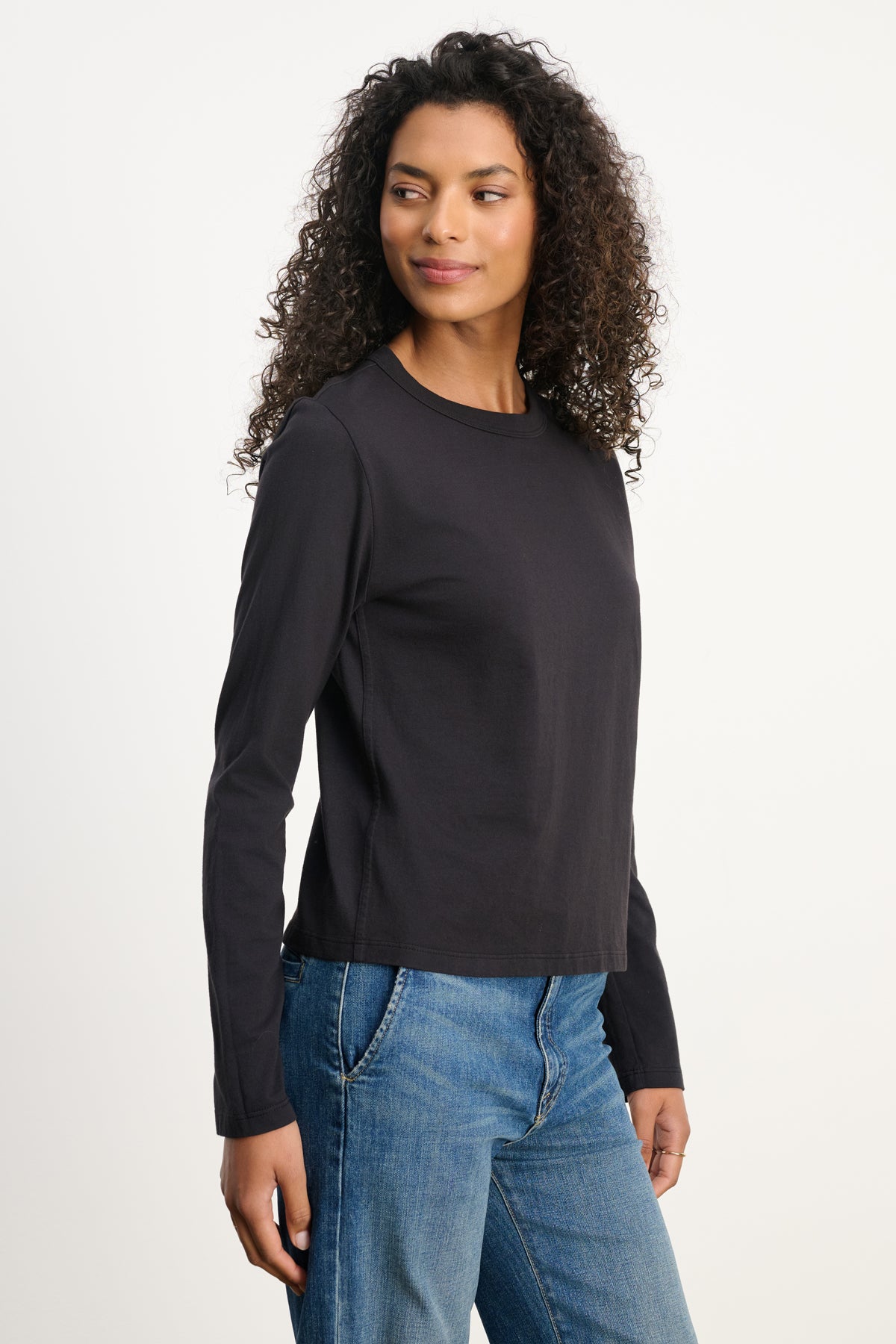 A woman with curly hair wears the SUSANNA TEE by Velvet by Graham & Spencer—a vintage-inspired, long-sleeved black cotton jersey shirt—paired with blue jeans, standing against a plain white background and looking slightly left.-39838373642433