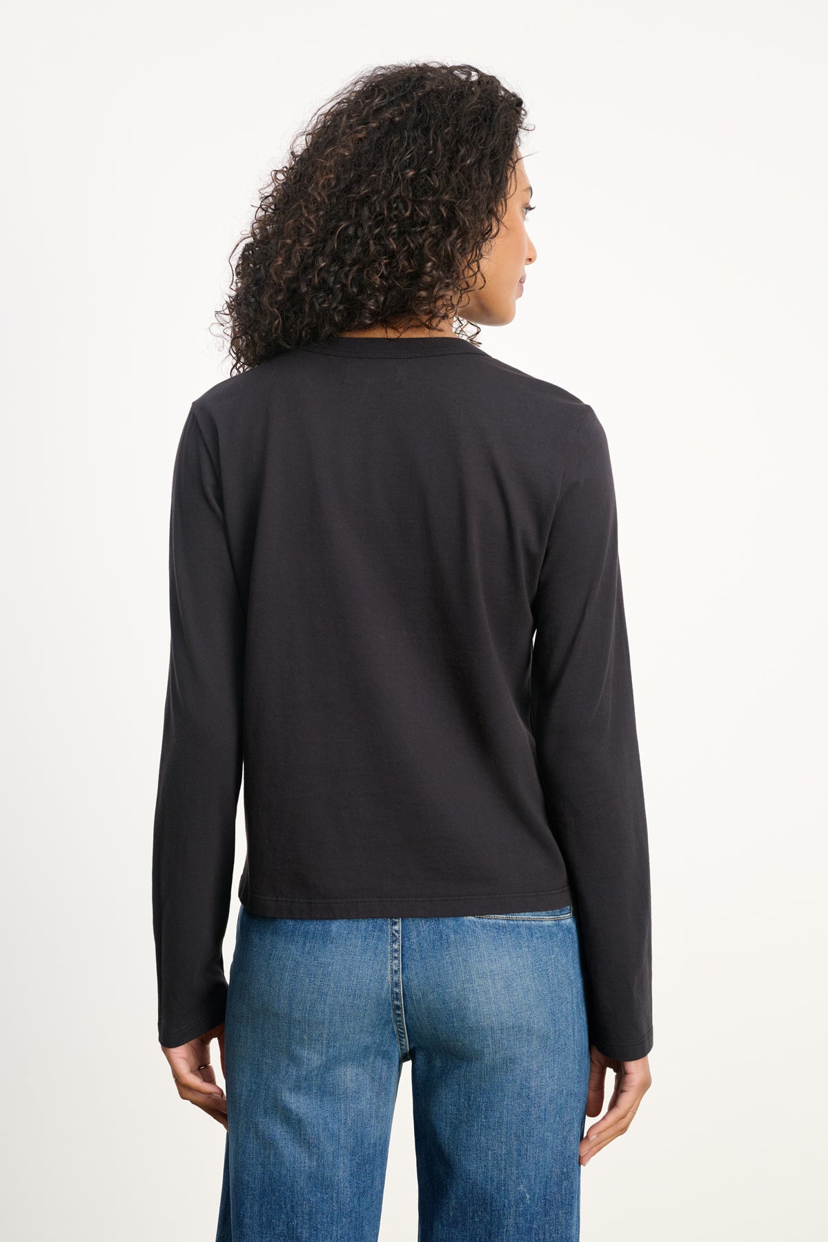 A person with curly hair models the SUSANNA TEE by Velvet by Graham & Spencer—a black, vintage-inspired long-sleeve shirt—paired with blue jeans, shown from the back against a plain background.-39838373609665