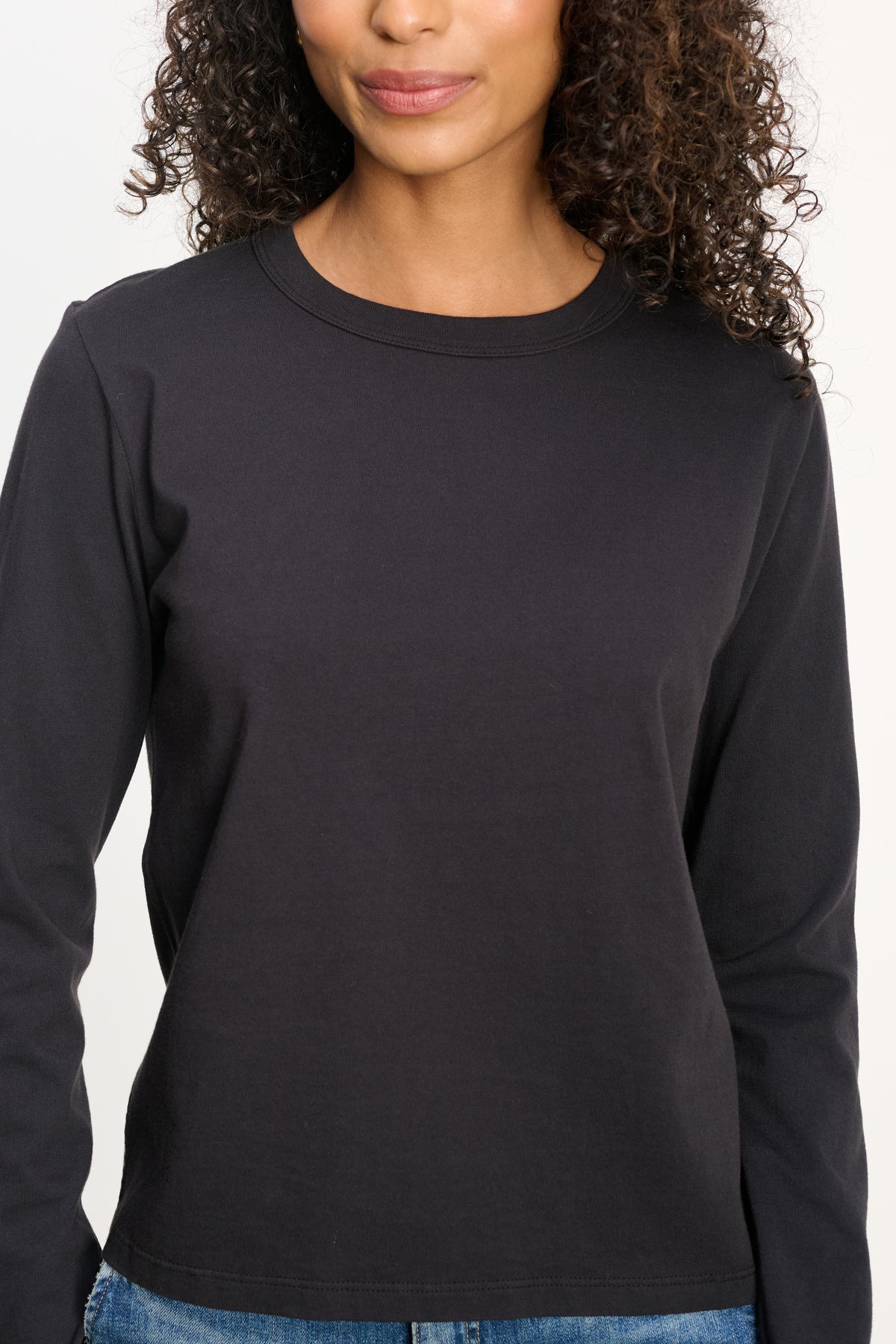 A woman with curly hair wears the SUSANNA TEE from Velvet by Graham & Spencer—a vintage-inspired, long-sleeve black cotton jersey top—paired with blue jeans, shown from shoulders to hips against a plain background.-39838373576897