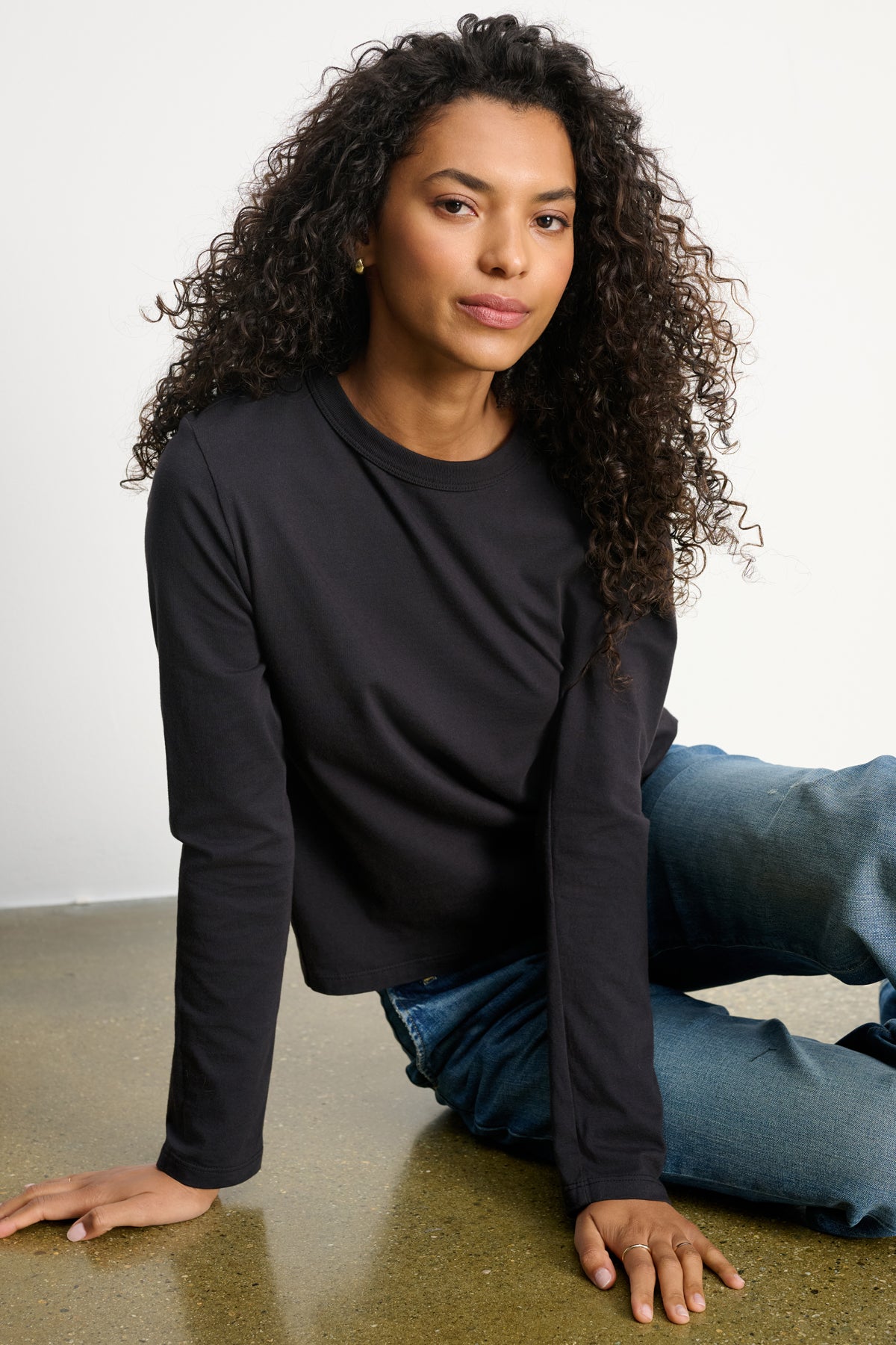 A woman with curly hair wears the Velvet by Graham & Spencer SUSANNA TEE, a vintage-inspired long-sleeve black cropped top, paired with blue jeans as she sits on the floor against a plain white background.-39838373413057