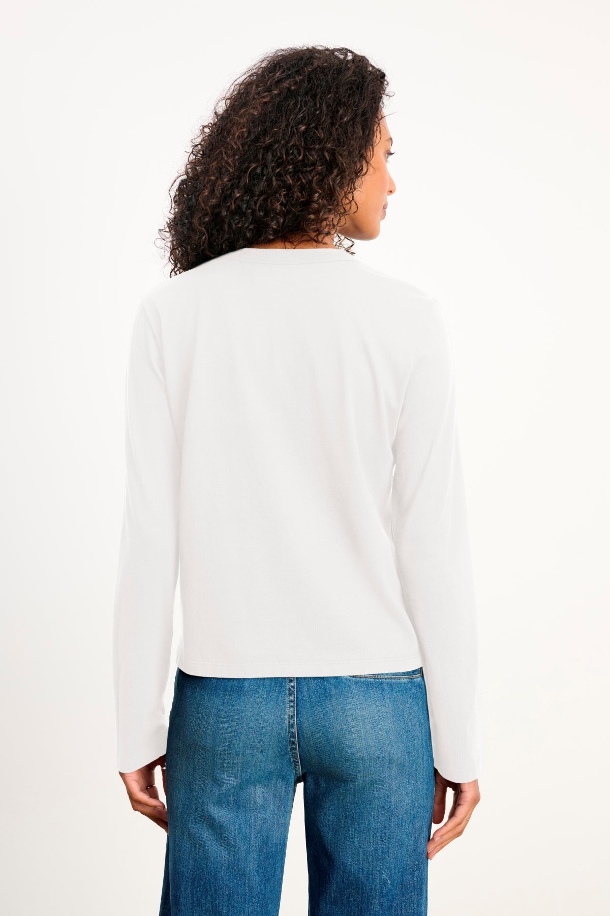A person with curly hair wears the SUSANNA TEE by Velvet by Graham & Spencer—a vintage-inspired long-sleeve white top—paired with blue jeans, standing with their back to the camera against a plain background.-39838401659073