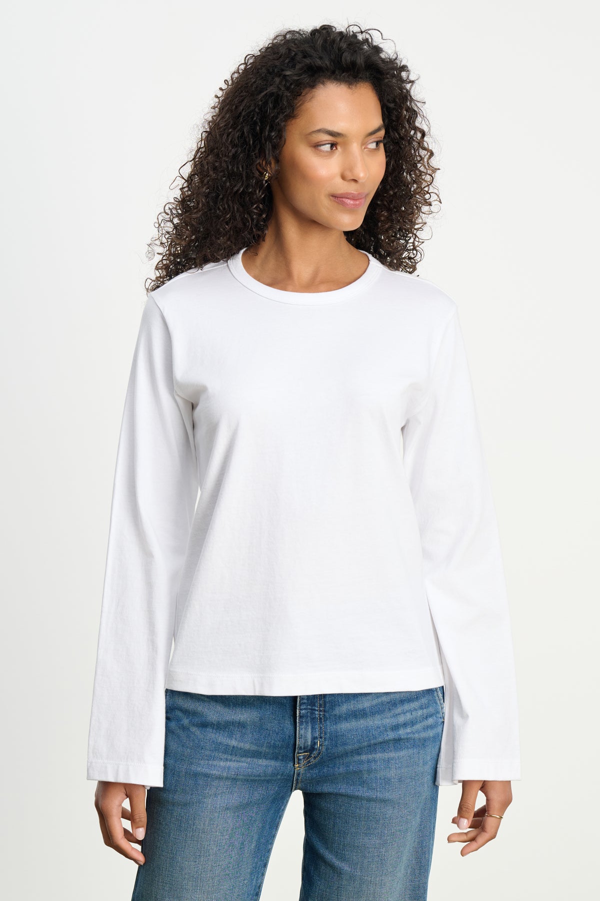 A person models the VEE TEE by Velvet by Graham & Spencer—a plain white long-sleeve shirt with a classic crew neckline—paired with blue jeans, standing against a white background and gazing slightly to the side.-39742396727489