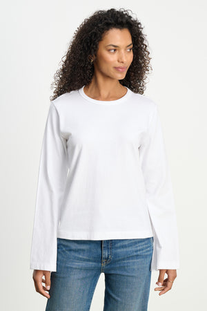 A person models the VEE TEE by Velvet by Graham & Spencer—a plain white long-sleeve shirt with a classic crew neckline—paired with blue jeans, standing against a white background and gazing slightly to the side.