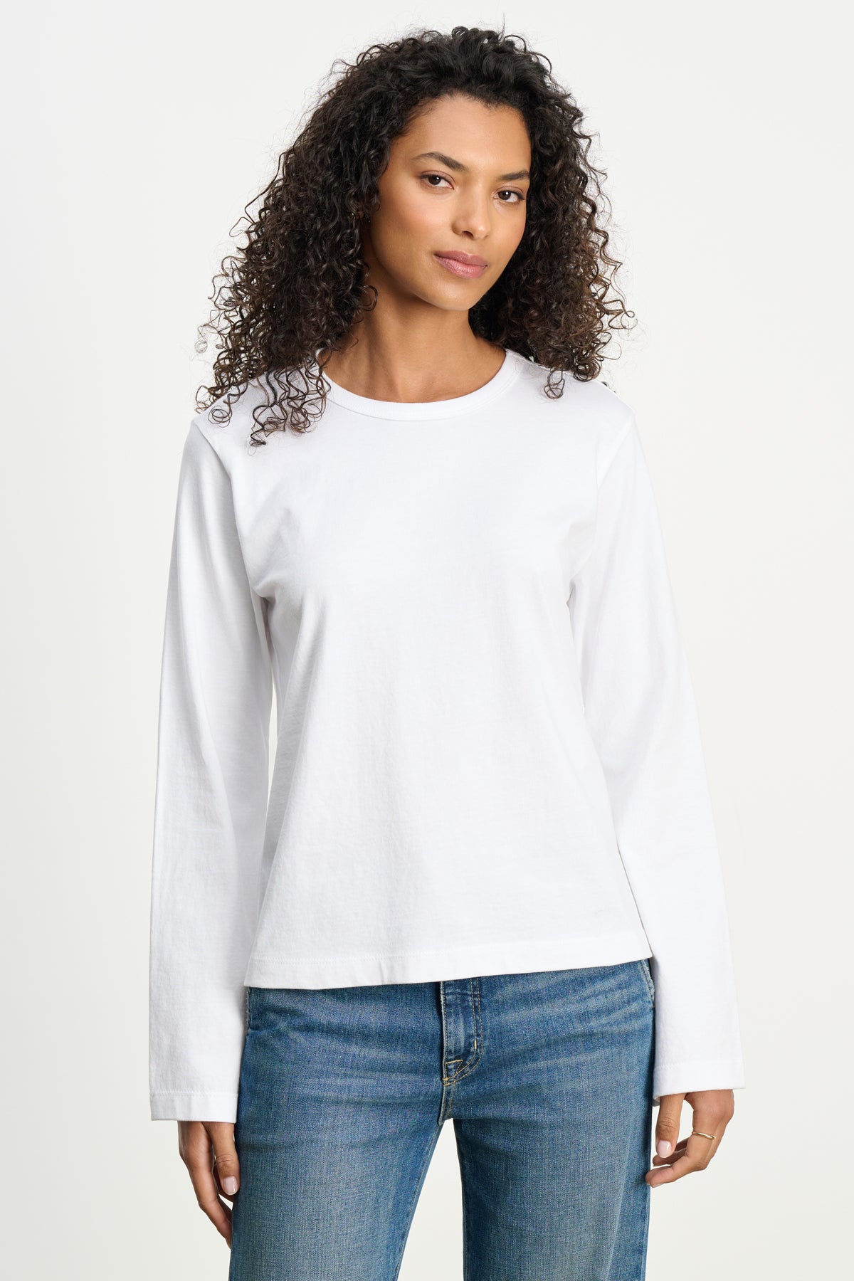 A woman with curly hair stands facing forward, wearing the VEE TEE by Velvet by Graham & Spencer—a classic cotton jersey crewneck in a boxy fit—paired with blue jeans against a neutral background.-39742396694721