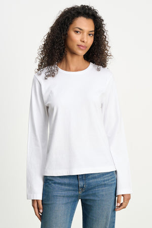 A woman with curly hair stands facing forward, wearing the VEE TEE by Velvet by Graham & Spencer—a classic cotton jersey crewneck in a boxy fit—paired with blue jeans against a neutral background.