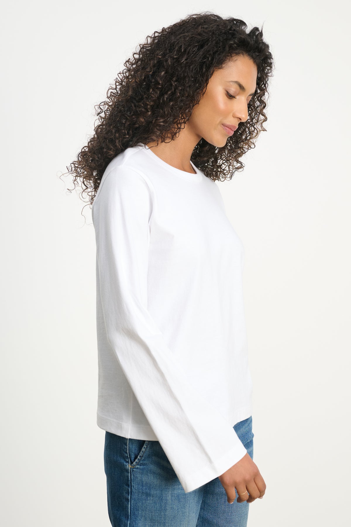 A woman with curly hair wears the VEE TEE by Velvet by Graham & Spencer—a long-sleeved white crewneck shirt—paired with blue jeans, standing in profile against a plain light background.-39742396629185