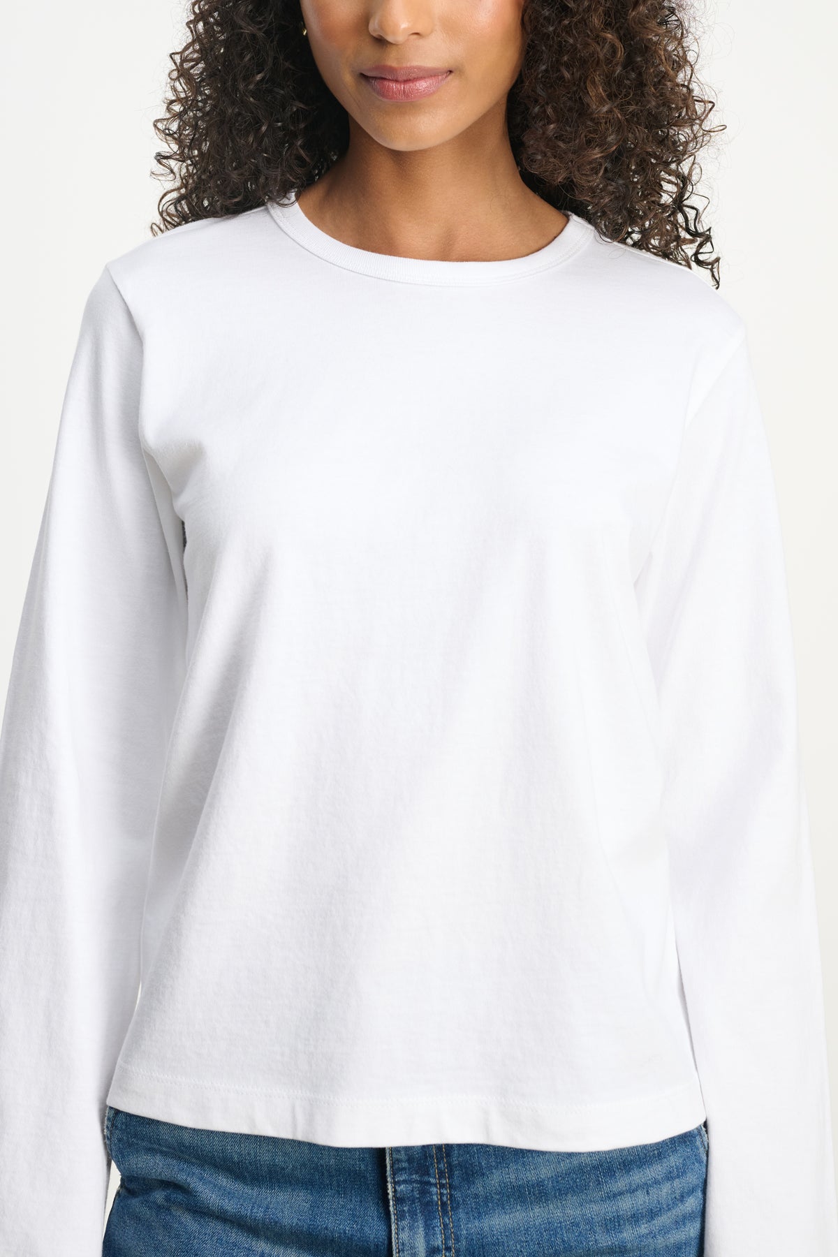 A person is shown from shoulders to hips against a neutral background wearing Velvet by Graham & Spencer's VEE TEE, a plain white long-sleeve shirt with a classic crew neckline, paired with blue jeans.-39742396596417