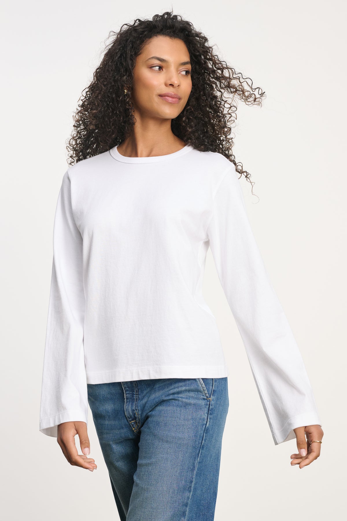 A person wears the VEE TEE long-sleeve white shirt by Velvet by Graham & Spencer with blue jeans, standing against a plain light background and looking slightly to the side.-39742396563649