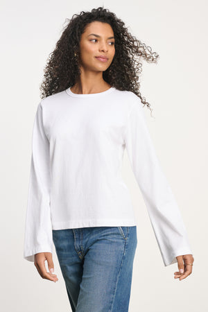 A person wears the VEE TEE long-sleeve white shirt by Velvet by Graham & Spencer with blue jeans, standing against a plain light background and looking slightly to the side.