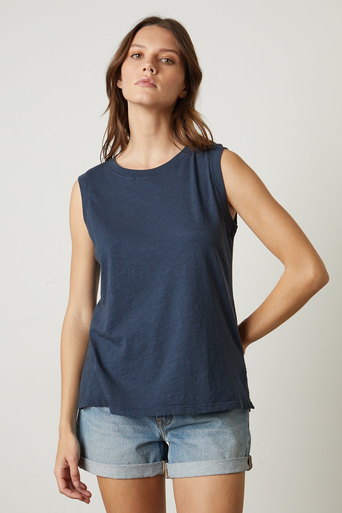   A person stands against a plain background, wearing an ELLEN VINTAGE SLUB TANK TOP by Velvet by Graham & Spencer and denim shorts. They have shoulder-length hair and a neutral expression. 