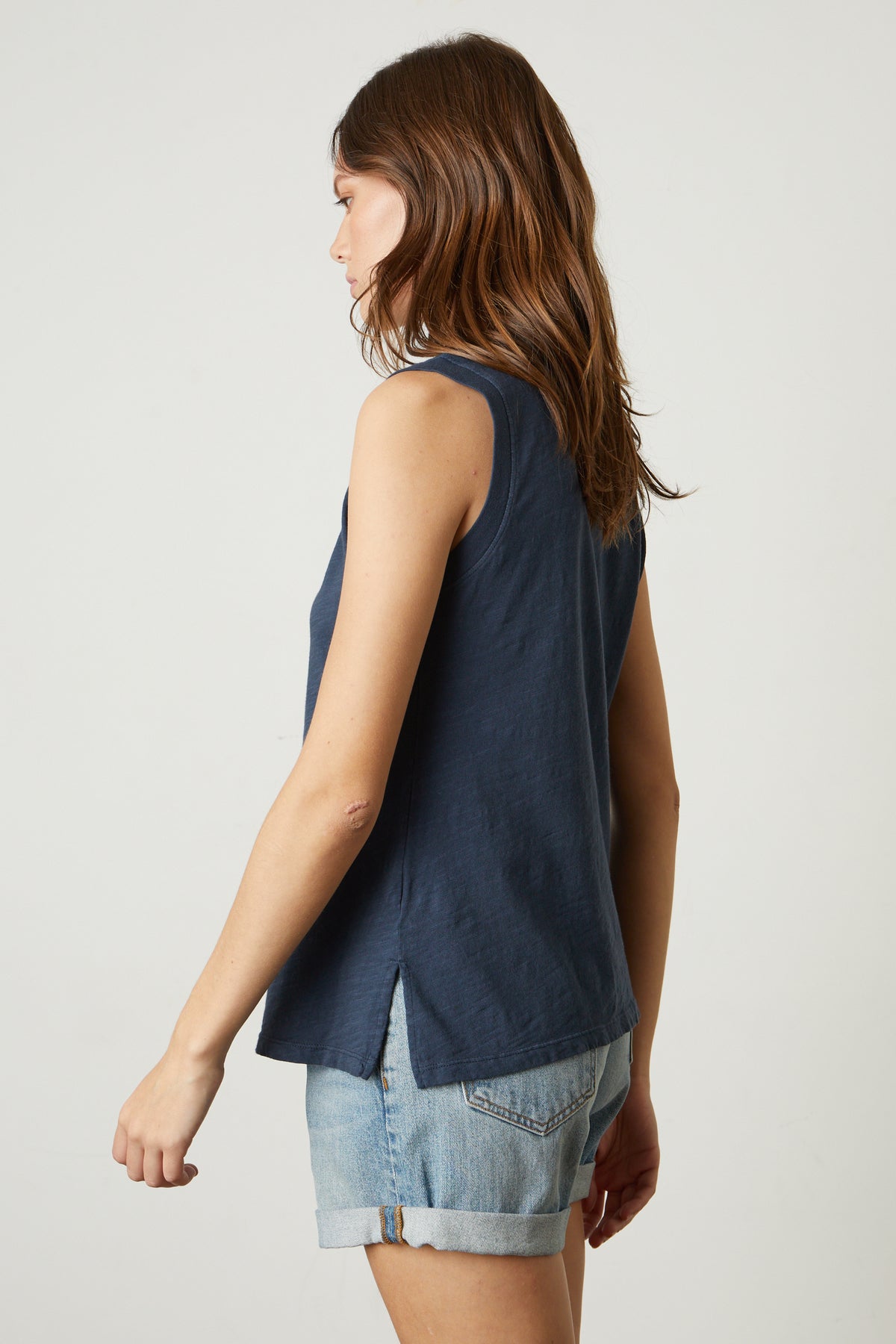 A woman with long brown hair, wearing a sleeveless navy top and denim shorts, embodies a comfortable-to-wear, classic style tank as she is seen from the side against a plain background in the ELLEN VINTAGE SLUB TANK TOP by Velvet by Graham & Spencer.-37249958412481