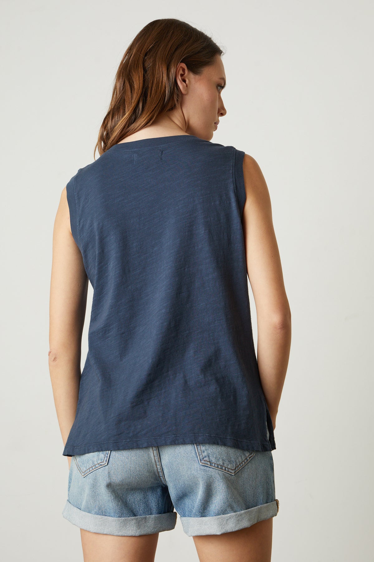   A woman is shown from the back, wearing a navy blue sleeveless top and denim shorts. Her brown hair is parted in the middle and falls past her shoulders. The background is plain and light-colored. The ELLEN VINTAGE SLUB TANK TOP by Velvet by Graham & Spencer gives off a comfortable, timeless vibe. 