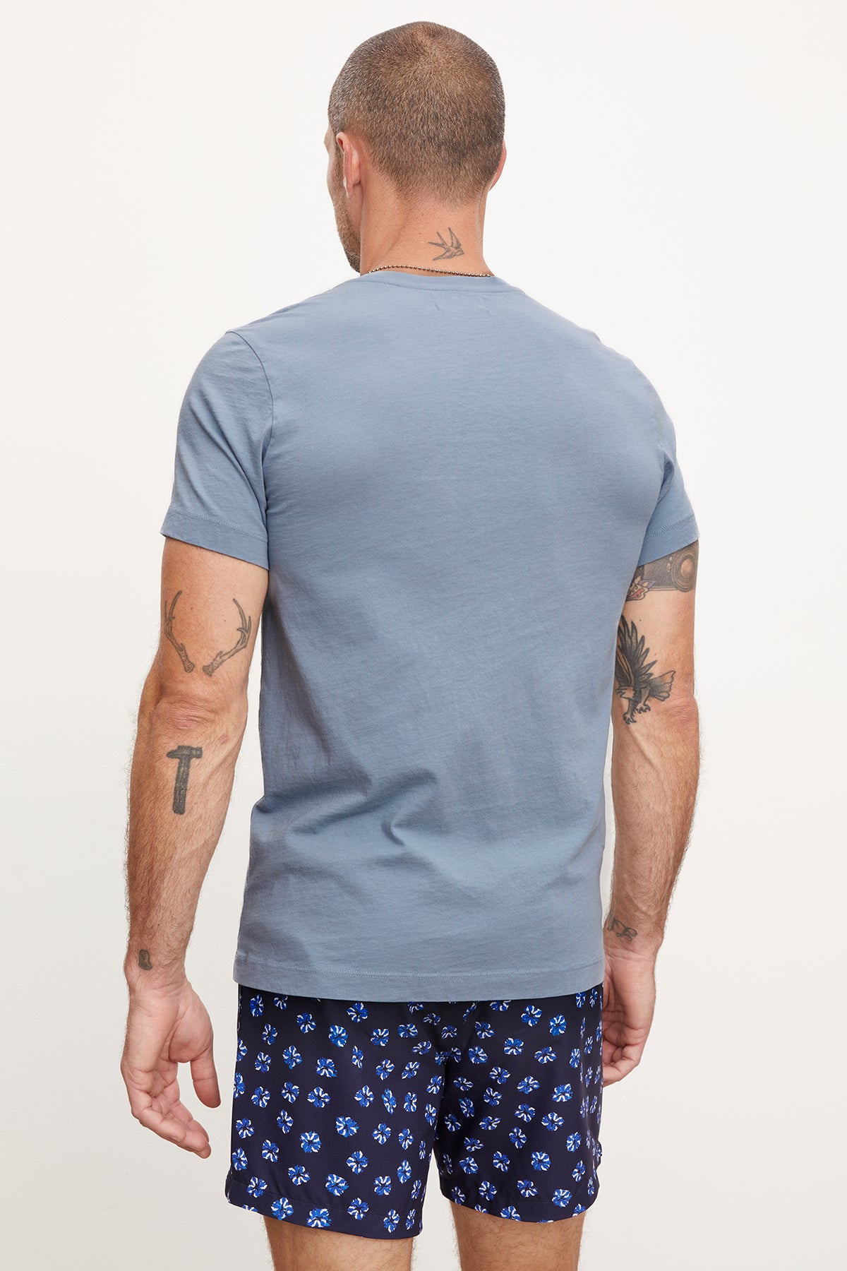   Rear view of a man wearing a gray Velvet by Graham & Spencer MARSHALL V-NECK TEE with patterned blue shorts, displaying visible tattoos on his arms and neck. 