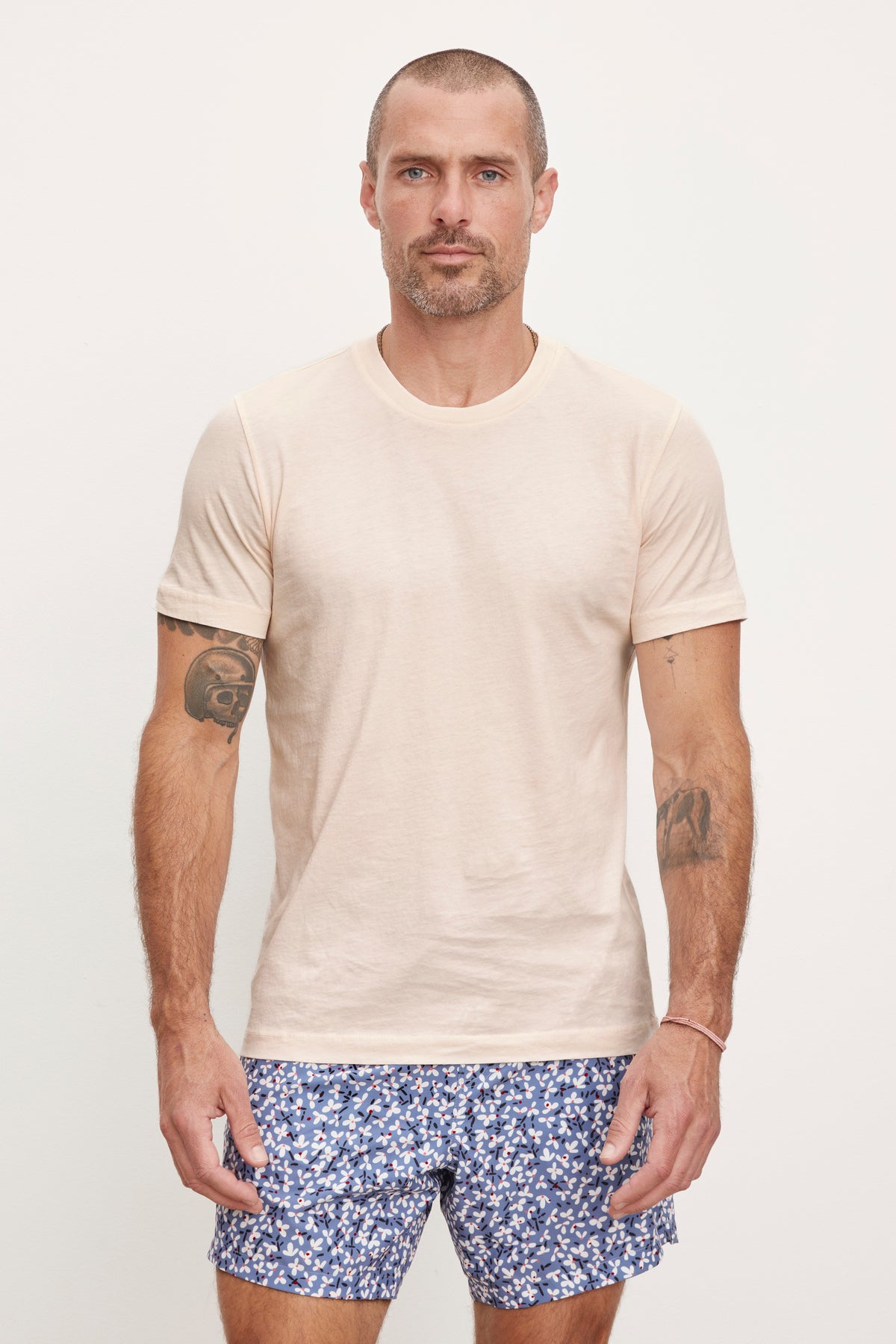   A man with tattoos on his arms wearing a Velvet by Graham & Spencer RANDY CREW NECK TEE and patterned blue shorts against a plain background. 