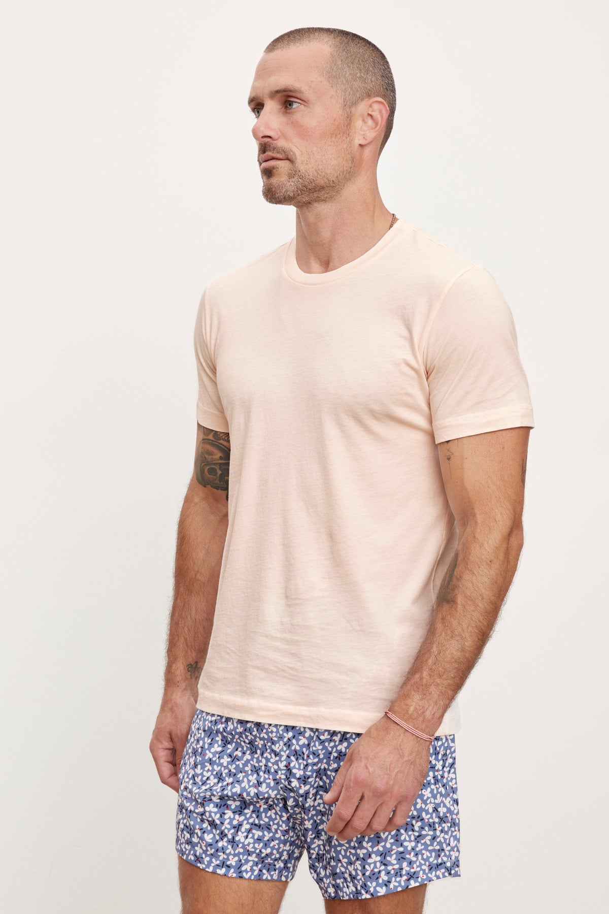 A man in a beige, 100% cotton Velvet by Graham & Spencer RANDY CREW NECK TEE and blue floral shorts standing against a white background, looking to his left.-36753568530625