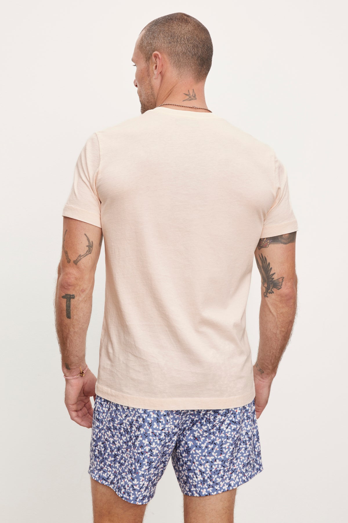 A man in a Velvet by Graham & Spencer RANDY CREW NECK TEE and blue patterned shorts stands facing away from the camera, showing visible tattoos on his arms and neck.-36753568563393
