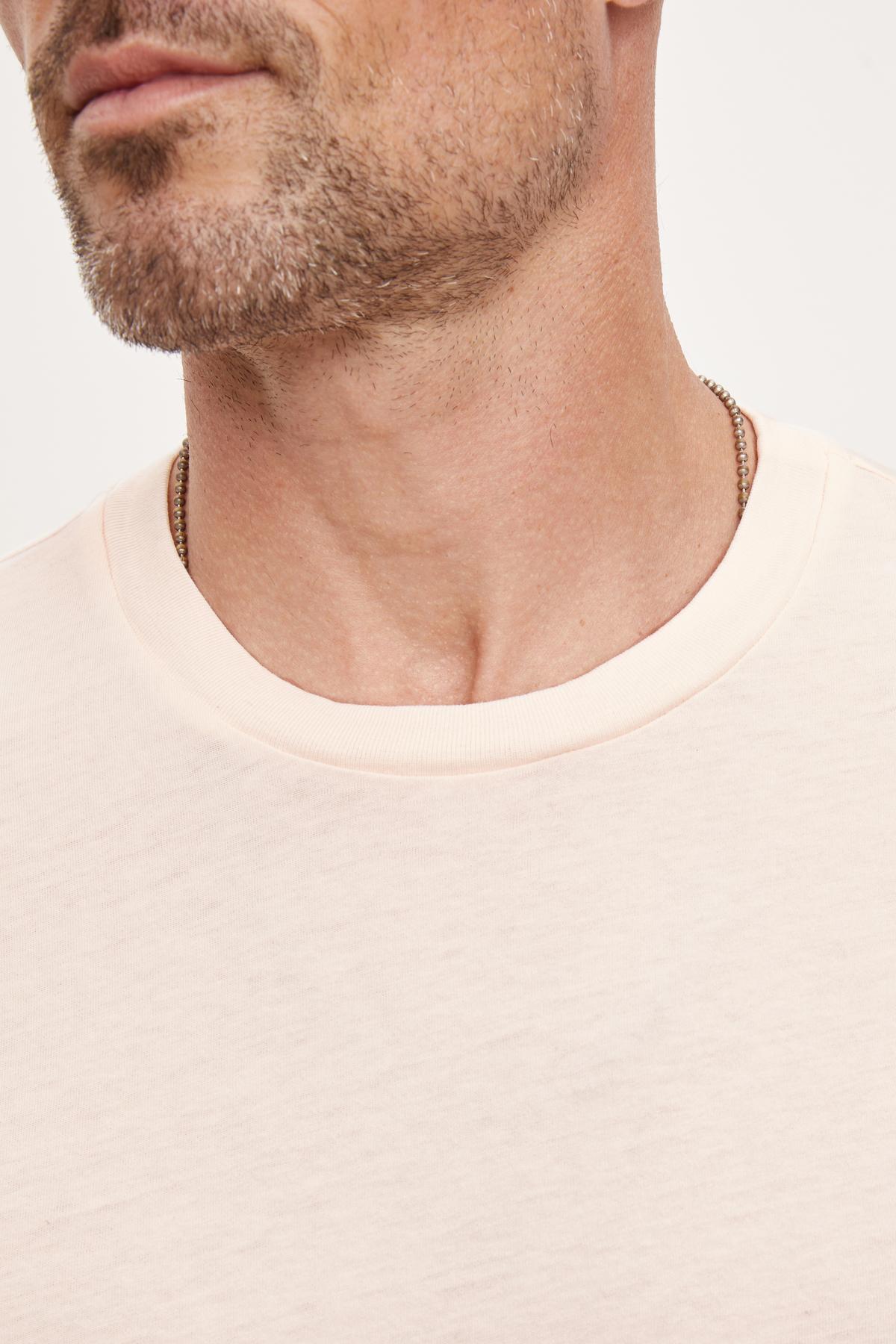   Close-up of a man wearing a Velvet by Graham & Spencer Randy Crew Neck Tee in light pink, made of 100% cotton, focusing on the neckline and the lower part of his face. 