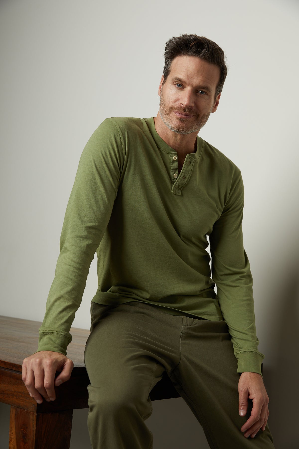 A man is sitting on a wooden table in a Velvet by Graham & Spencer BRADEN HENLEY shirt.-35567416770753