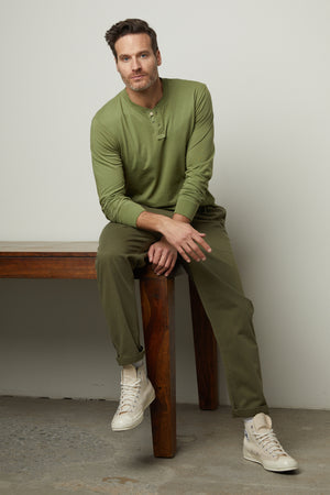 A man sitting on a wooden bench in a green Velvet by Graham & Spencer cotton woven henley shirt.
