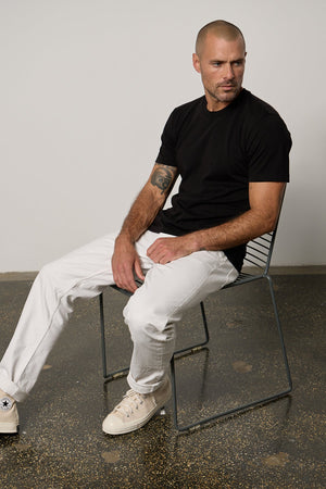 a man sitting on a chair wearing a Velvet by Graham & Spencer RANDY CREW NECK TEE and white jeans.